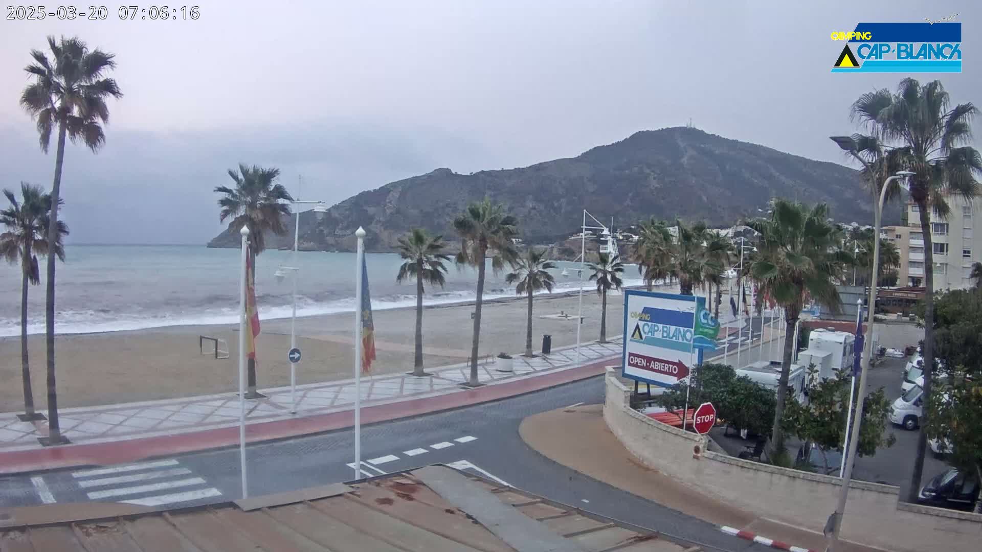A sandy beach with palm trees and a calm sea under an overcast sky is seen from an elevated position, showing a road and buildings in the background.