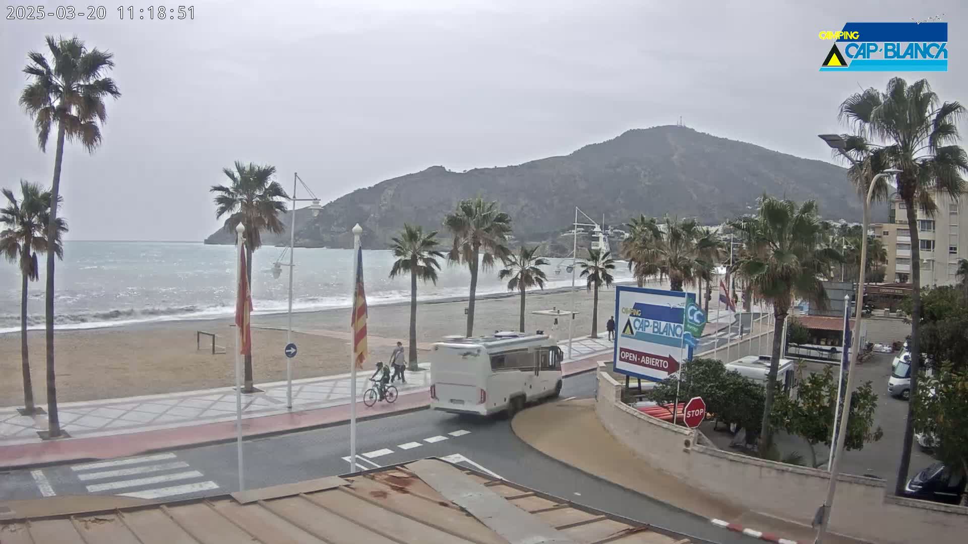 A beach scene on an overcast day shows a sandy beach with palm trees, a calm sea, a motorhome, and people riding bicycles along a paved walkway near a hill.