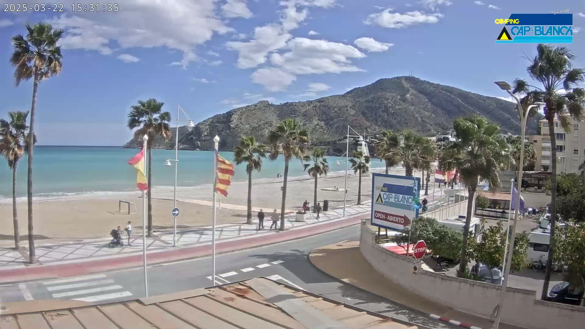 A sandy beach with palm trees and turquoise water is visible under a partly sunny sky, with a mountain in the background and a road leading to a parking area.