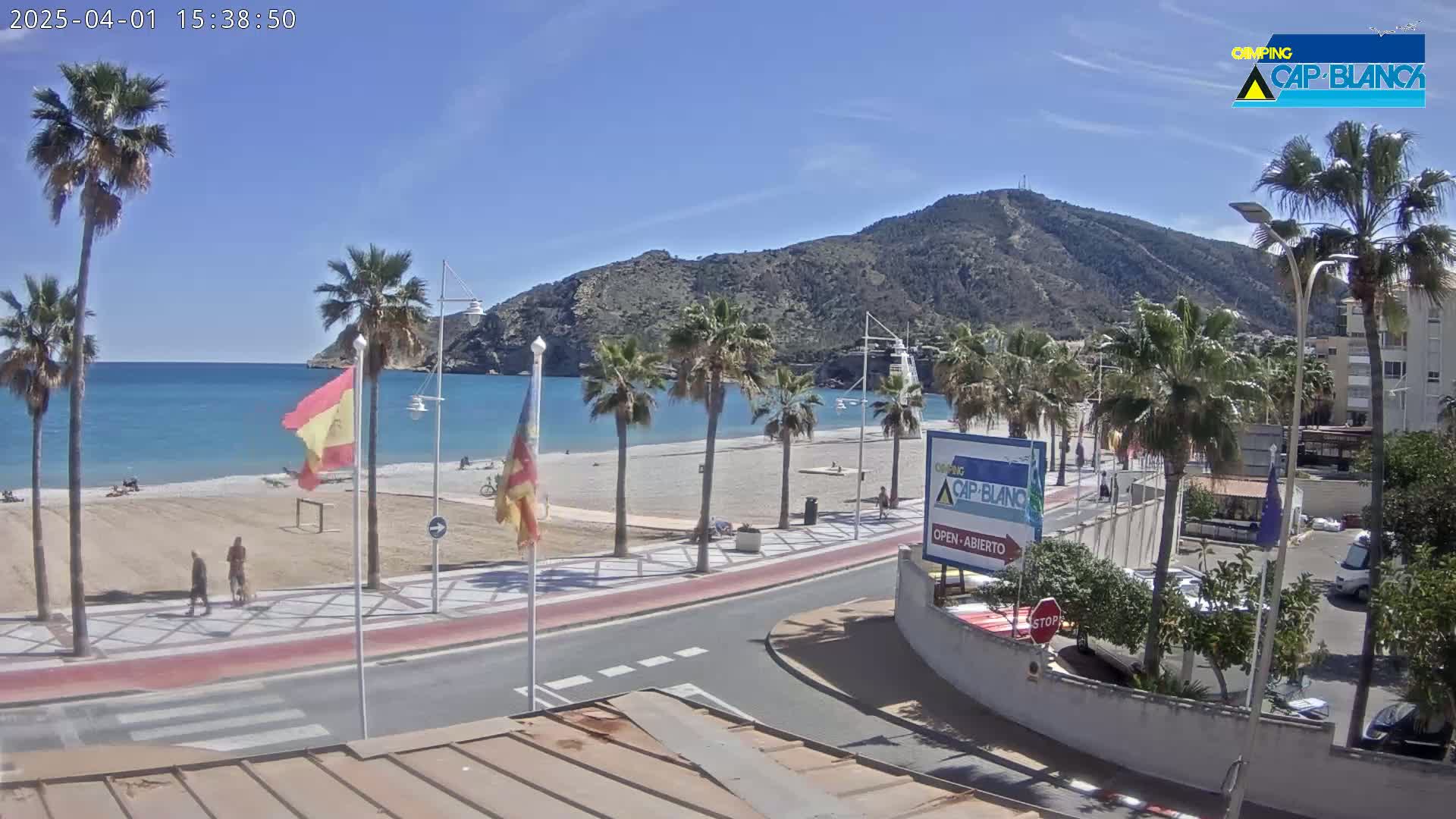A sandy beach with palm trees lines a paved walkway next to a calm, clear blue ocean, all backed by a brown mountain under a sunny sky.