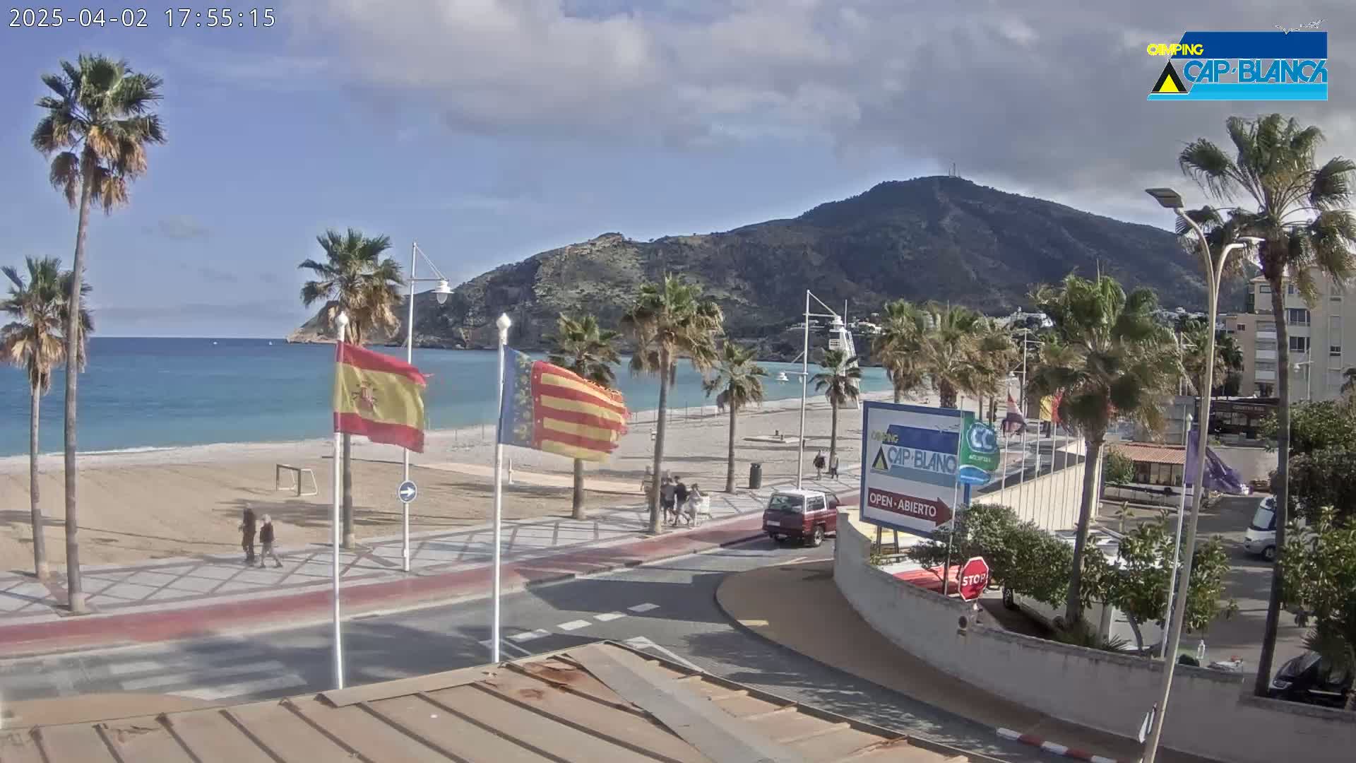 A sandy beach with palm trees and a calm ocean is visible beneath a partly cloudy sky, with a mountain in the background and a few people walking along the beachside road.