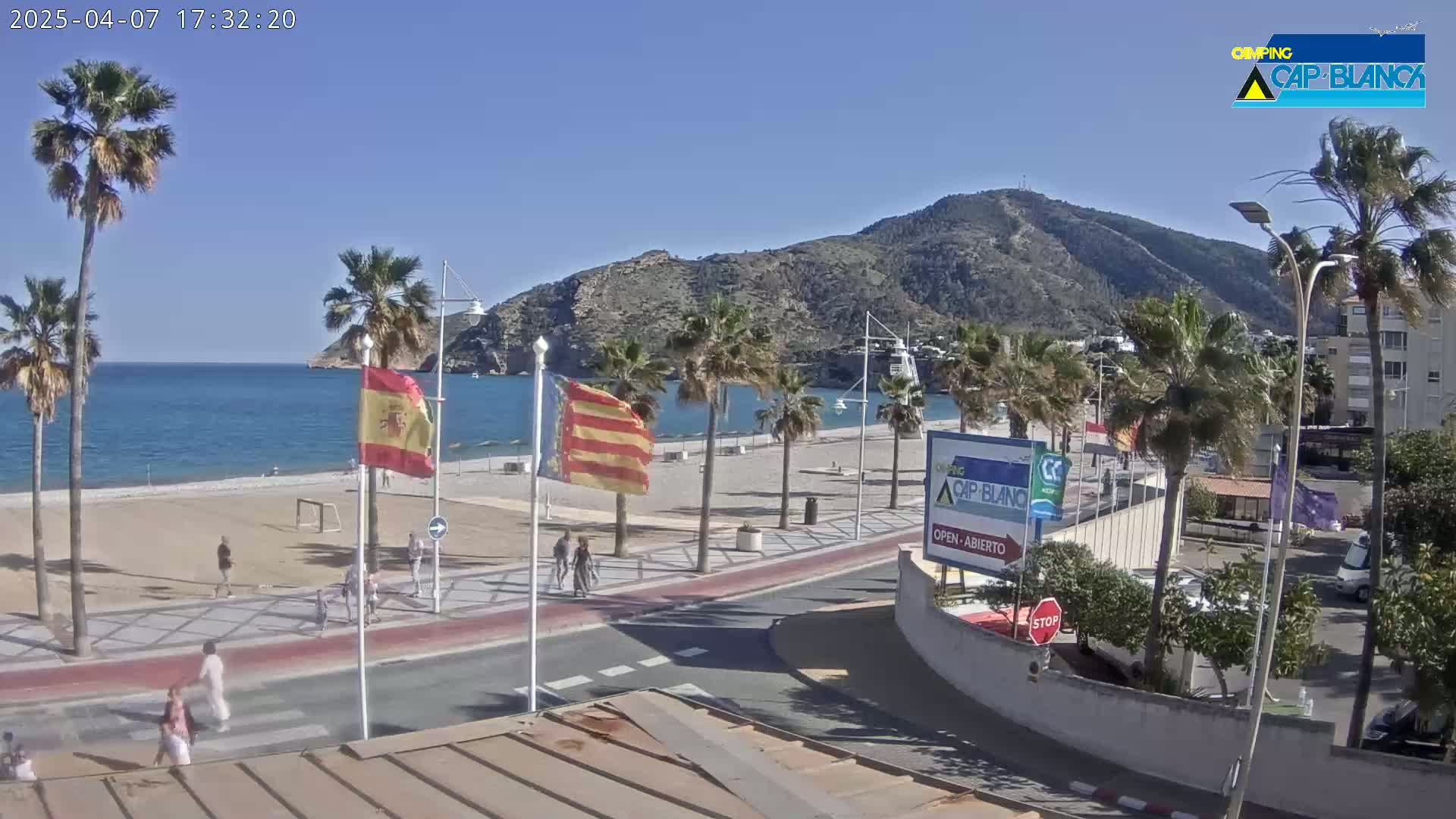 A sunny beach scene shows palm trees lining a paved walkway next to a sandy beach with a calm sea and mountains in the background.