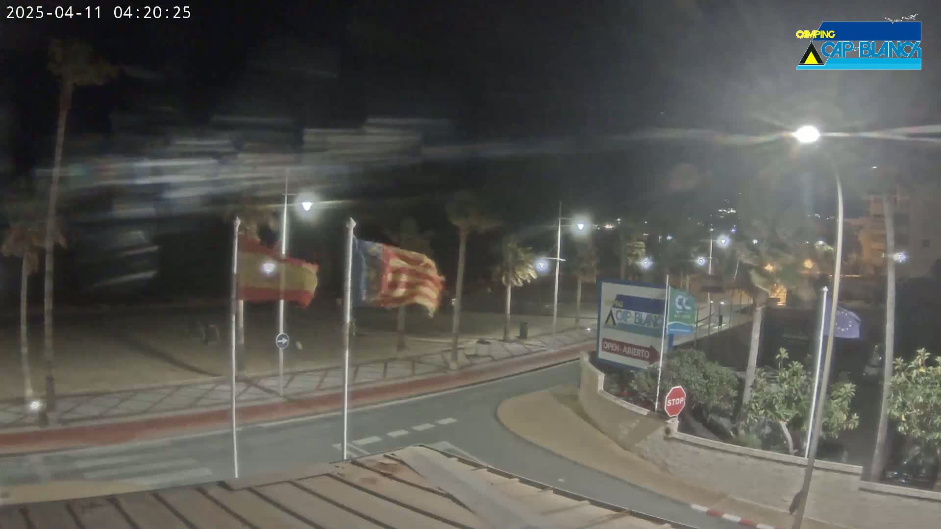 A nighttime, windy scene shows a roadway curving near a beach with palm trees, Spanish and Catalan flags waving, and streetlights illuminating the area.