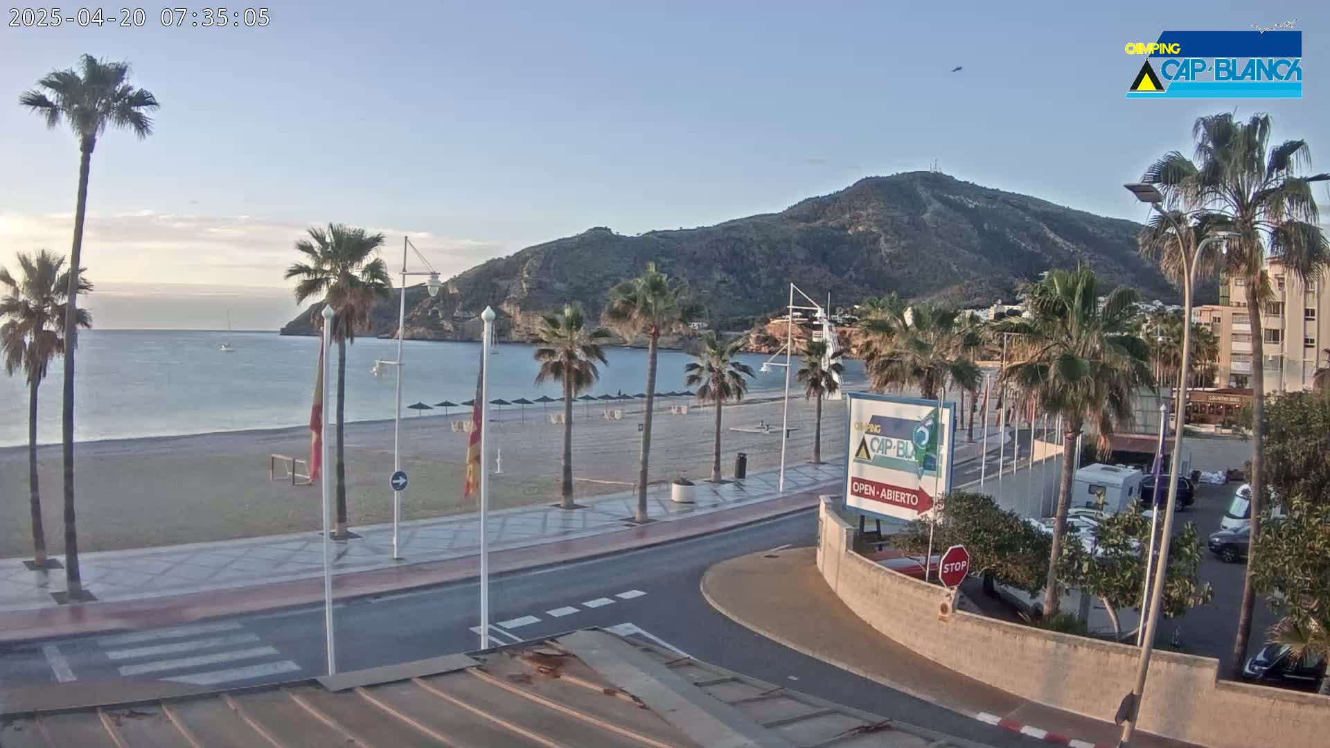 A mostly clear, early morning view of a beach with palm trees, a calm sea, and a large hill in the background; a road leads from the beach to a parking area.
