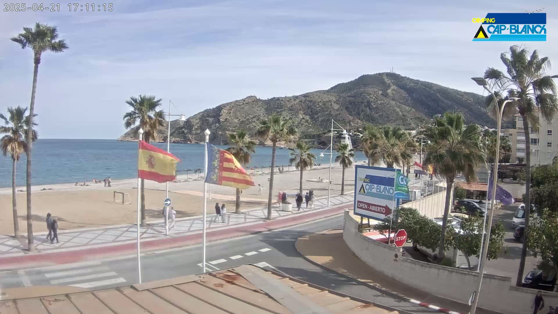 A sunny day at a beachside promenade, featuring palm trees, a sandy beach with a few people, and a mountain in the background.