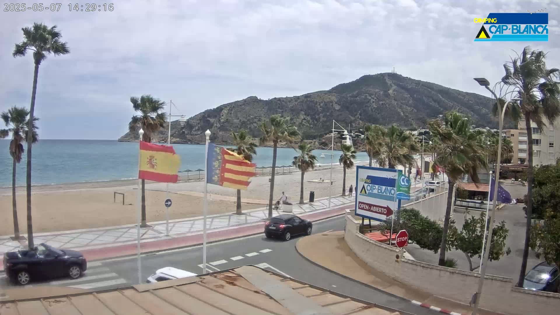 A beachside road with palm trees, a sandy beach, calm blue water, and a mountain in the background under partly cloudy skies.