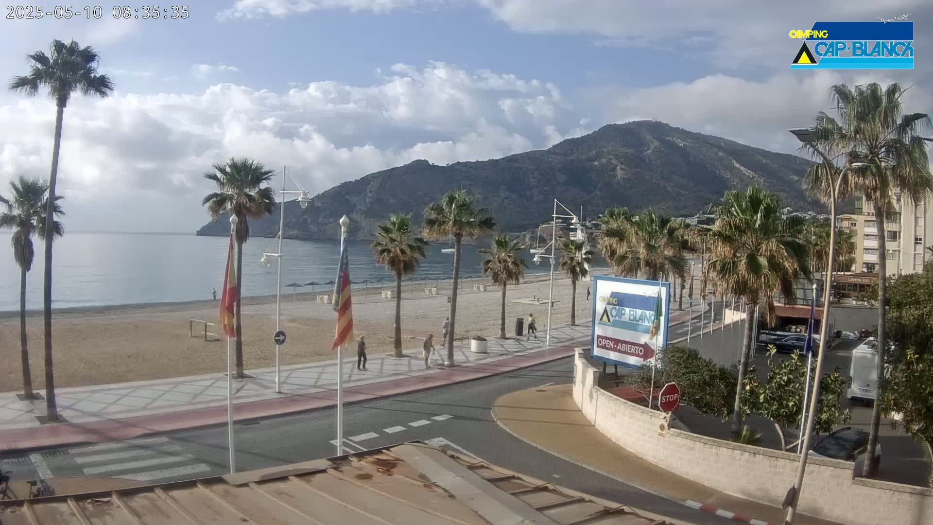 A partly cloudy day at a beachside promenade, featuring palm trees, a sandy beach, calm ocean water, and a mountain in the background.