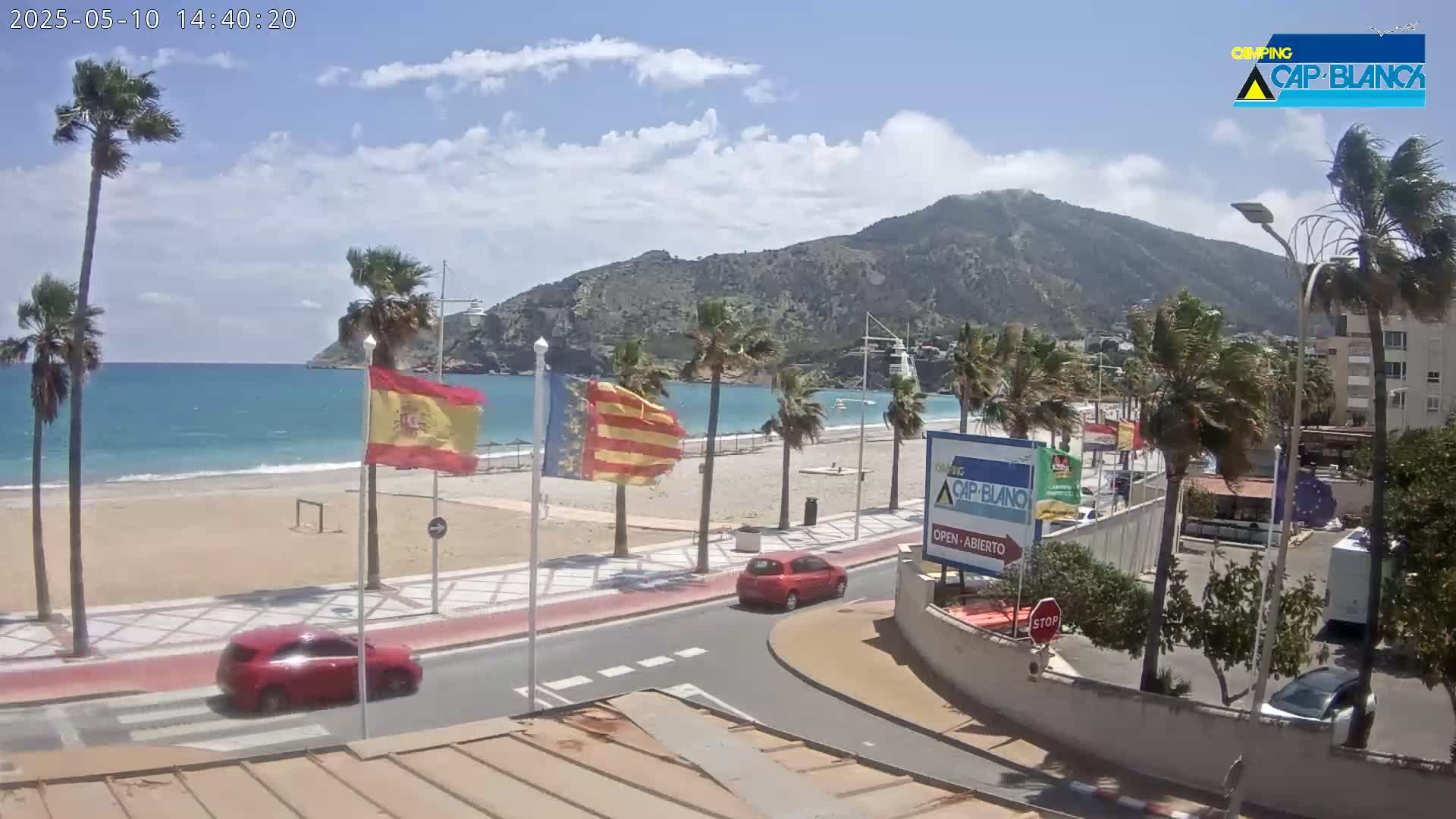 A sandy beach with calm, blue water is backed by a road lined with palm trees and buildings, all under a partly sunny sky.