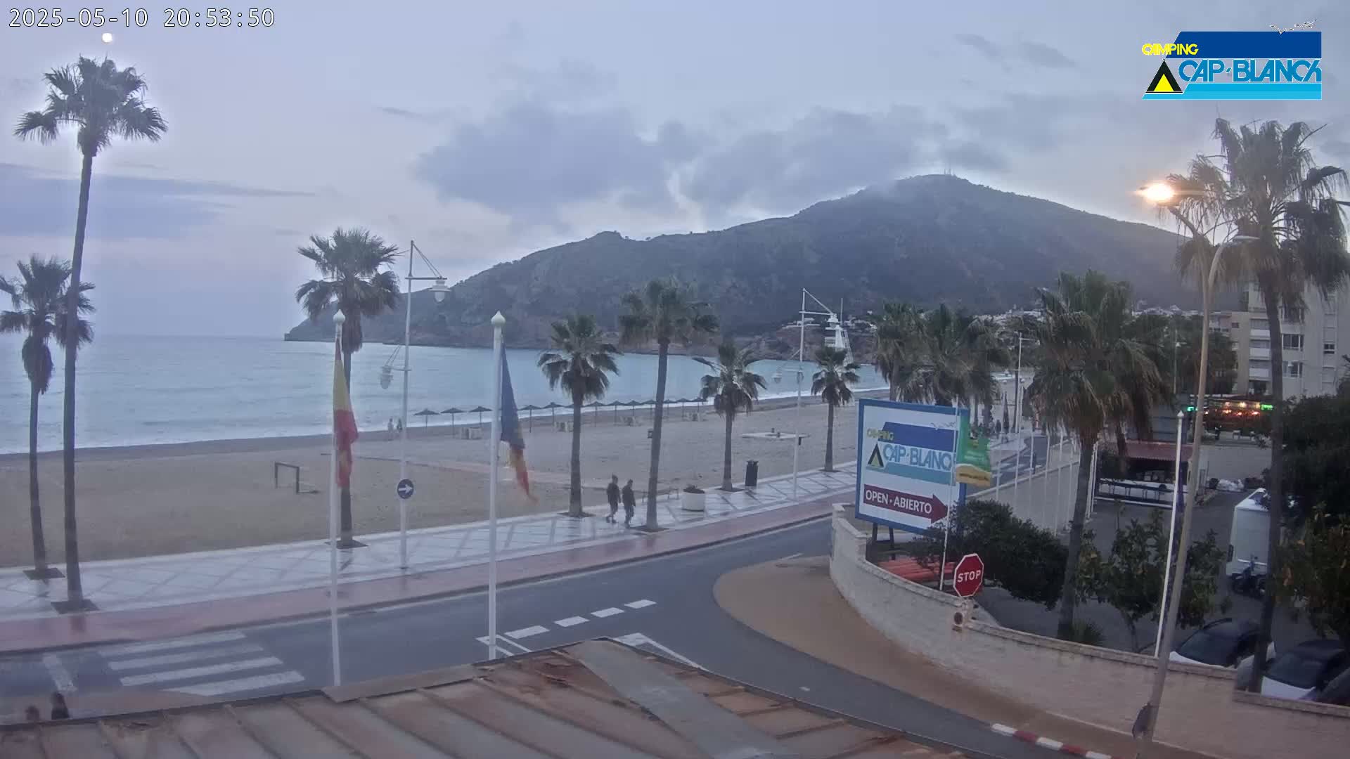A mostly cloudy evening scene shows a beach with palm trees, a calm sea, a mountain in the background, and a paved road leading to a parking area.