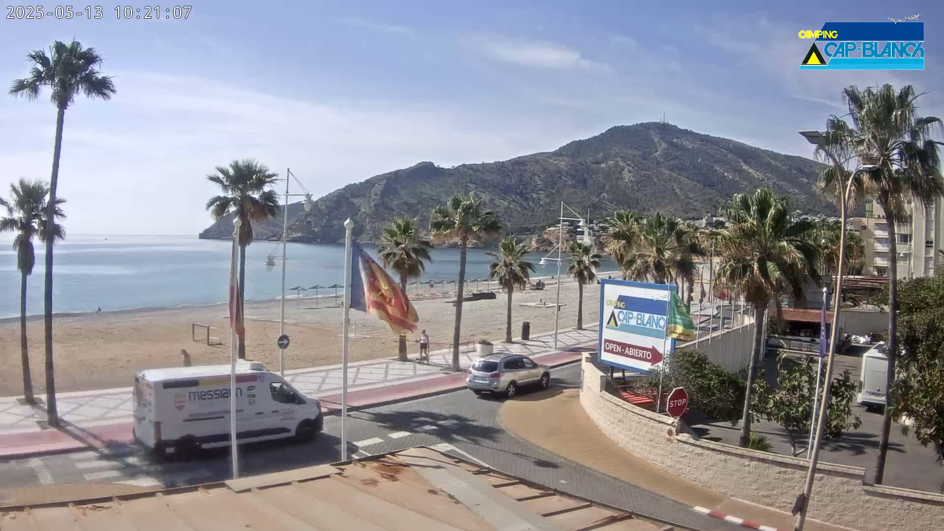 A sunny day at a beachside town shows a sandy beach, calm ocean, palm trees, parked cars, and a mountain in the background.
