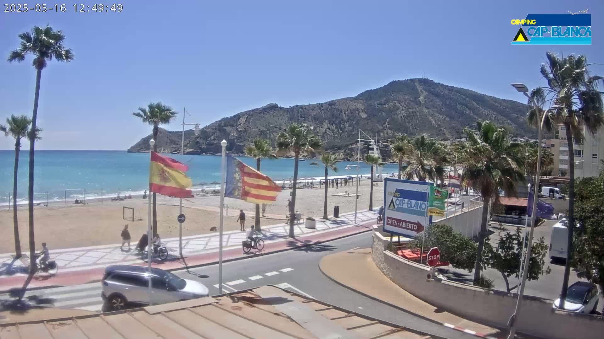 A sandy beach with calm, turquoise water is backed by a paved walkway, palm trees, and a mountain under a clear blue sky.
