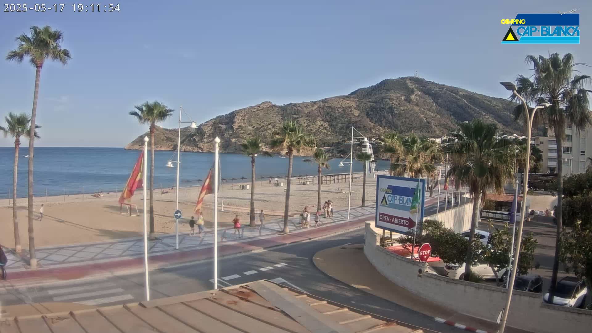 A sandy beach with palm trees and a few people is visible, next to a paved walkway and road, with a calm blue sea and a mountain in the background under a clear sky.