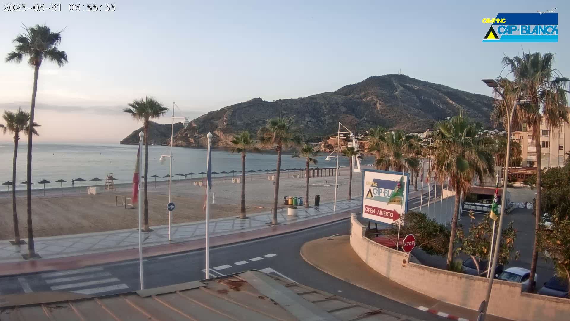 A mostly empty sandy beach with palm trees and a calm sea under a clear, light blue sky is viewed from a slight elevation, showing a road leading to it.