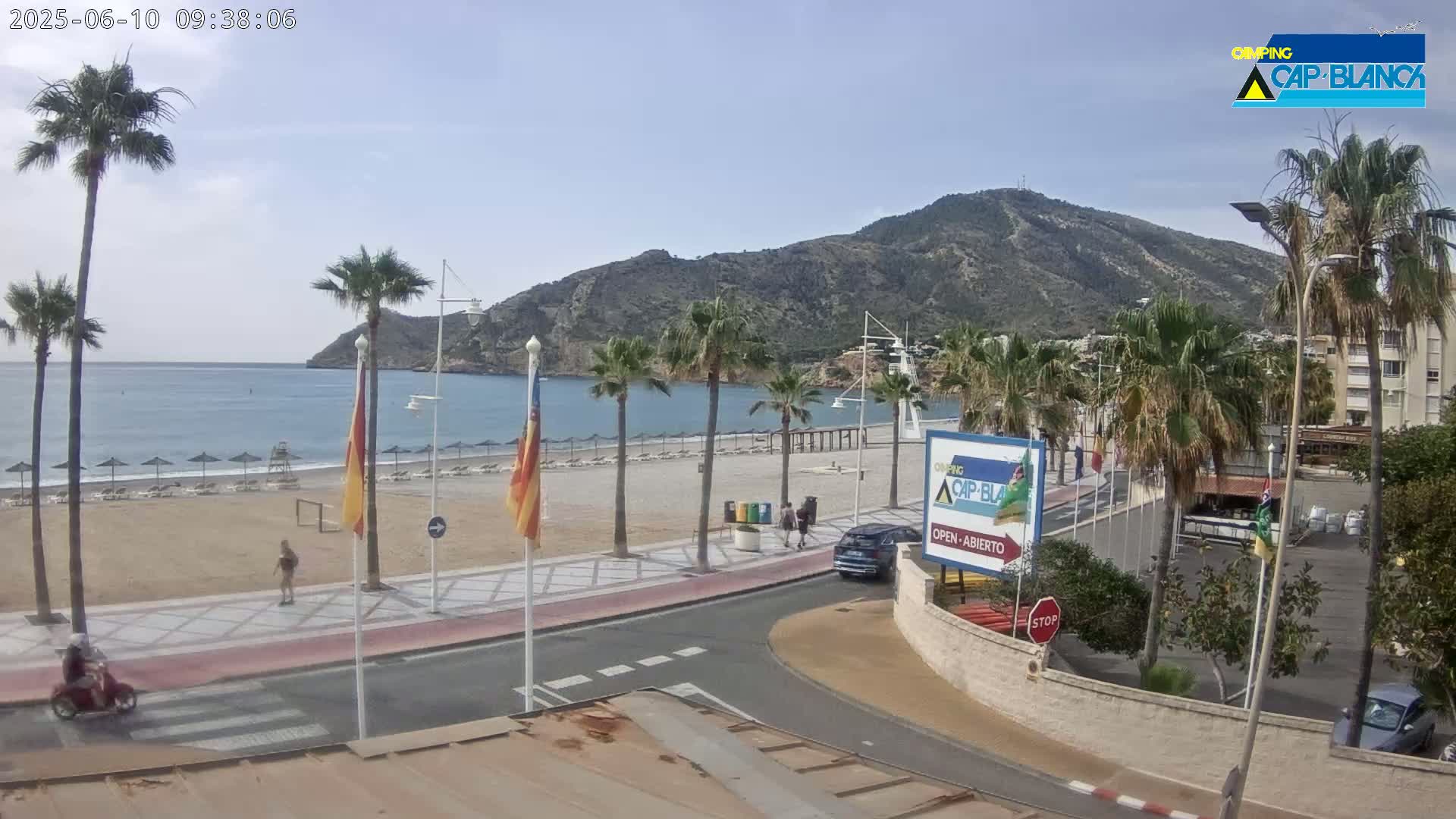 A mostly sunny day shows a sandy beach with palm trees, a calm ocean, and a mountainous backdrop next to a road with some vehicles and pedestrians.