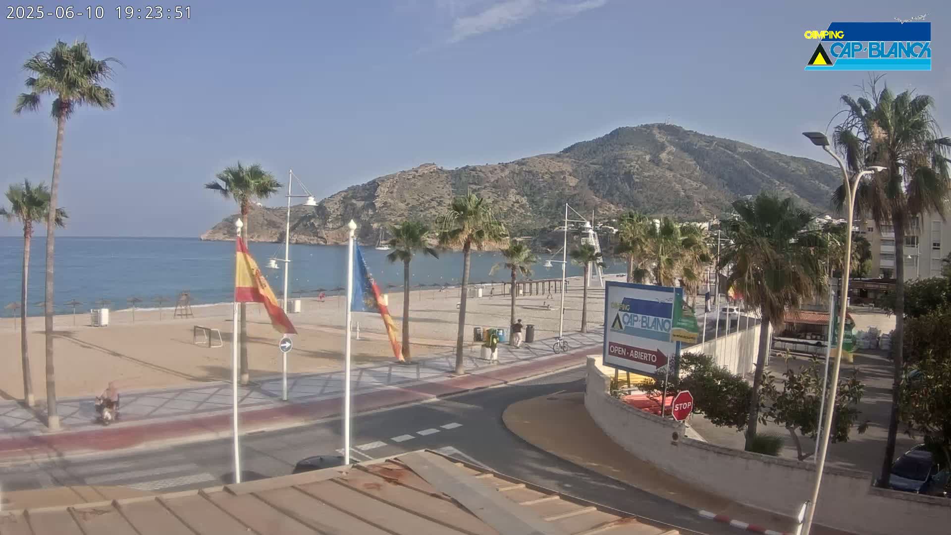 A sunny beach scene shows a sandy beach, calm blue water, palm trees, a mountain in the background, and a road with pedestrians and a bicycle.