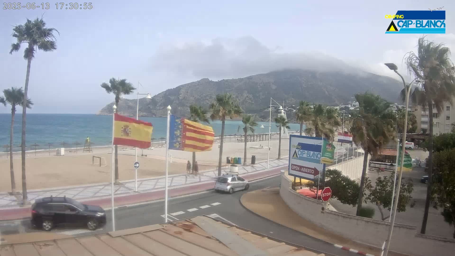 A mostly sunny beach scene with a hazy mountain in the background, palm trees lining a paved beachfront road, and several cars parked and driving.