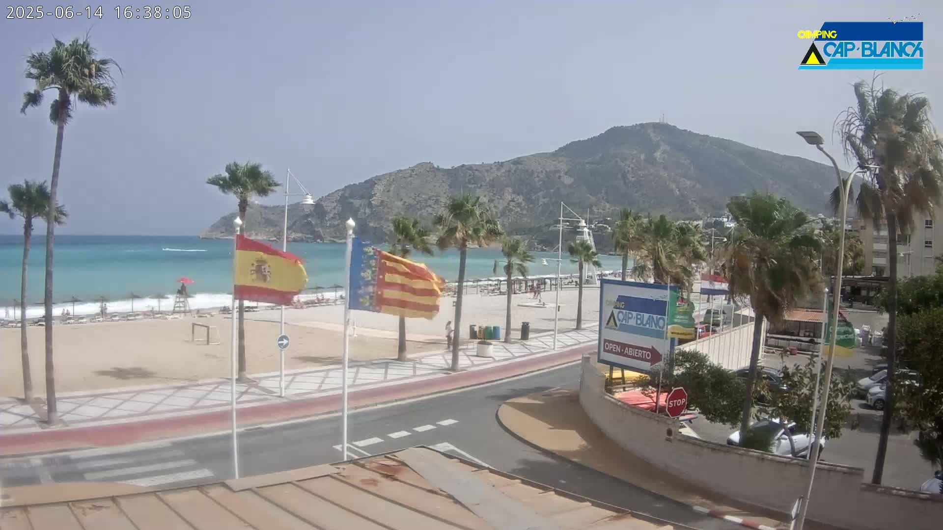 A sandy beach with turquoise water is visible beneath a mostly clear sky, backed by palm trees, a coastal road, buildings, and a mountain.