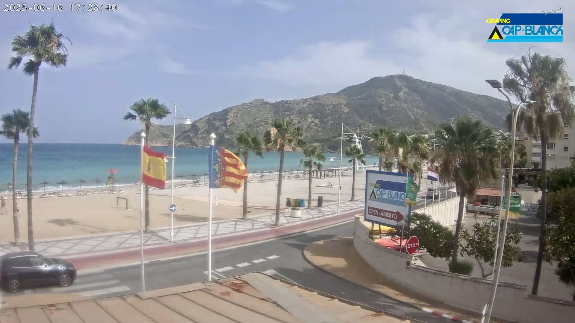 A sandy beach with palm trees and turquoise water is situated beside a road, with a mountainous backdrop under a mostly sunny sky.