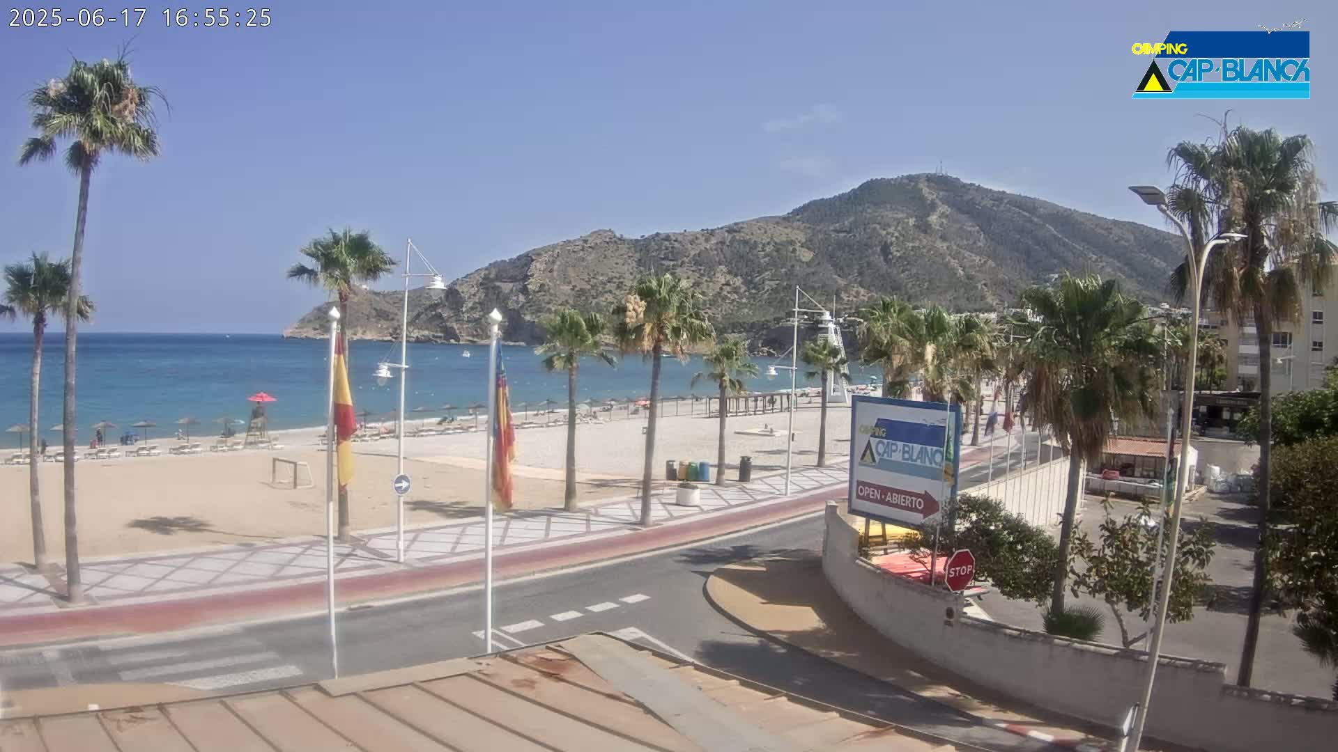 A sunny beach scene shows a sandy beach with people, palm trees, and a calm blue ocean, all set against a backdrop of a large mountain under a clear blue sky.