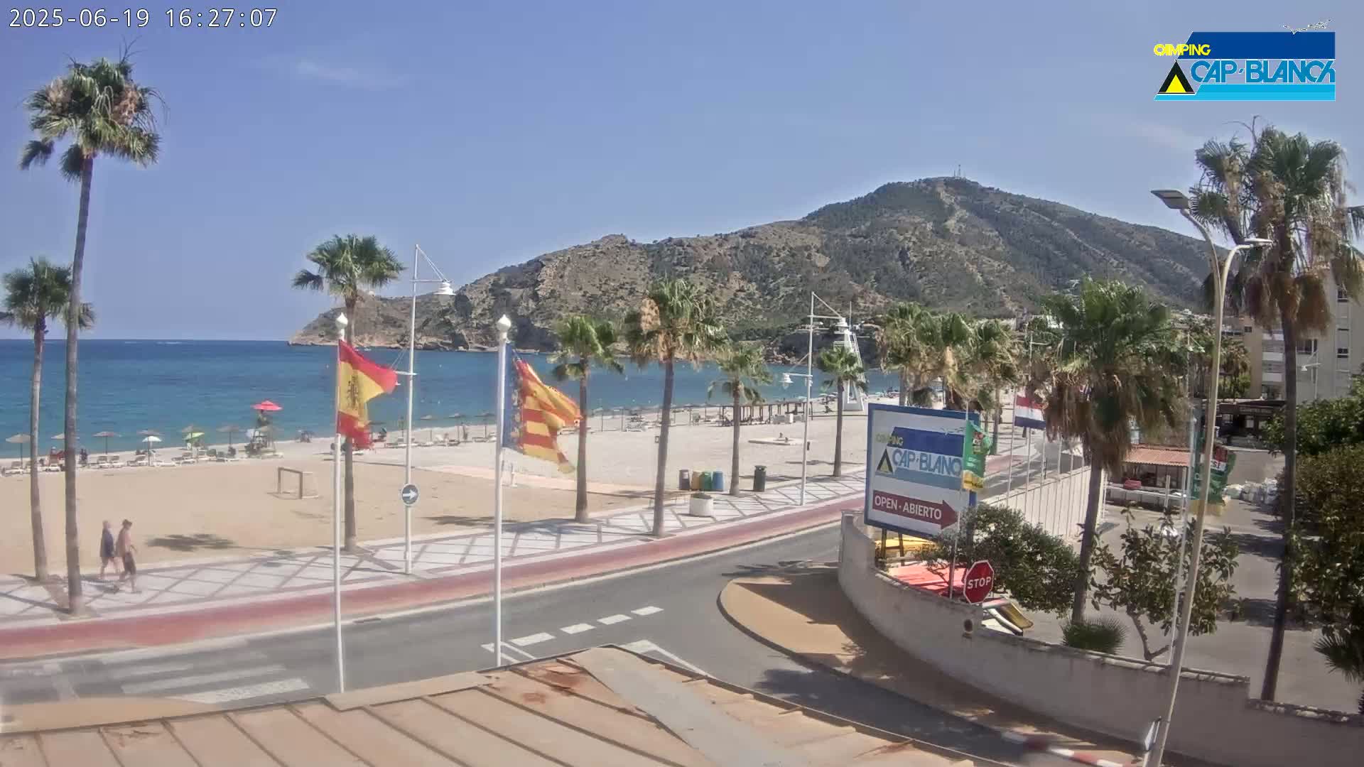 A sandy beach with palm trees and a few people on it is next to a calm, blue ocean and a rocky mountain under a clear, sunny sky.