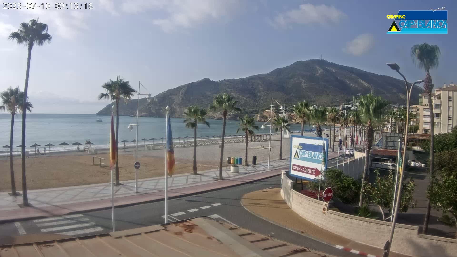 A sandy beach with calm water and palm trees lines a paved walkway leading to buildings under a mostly clear sky.