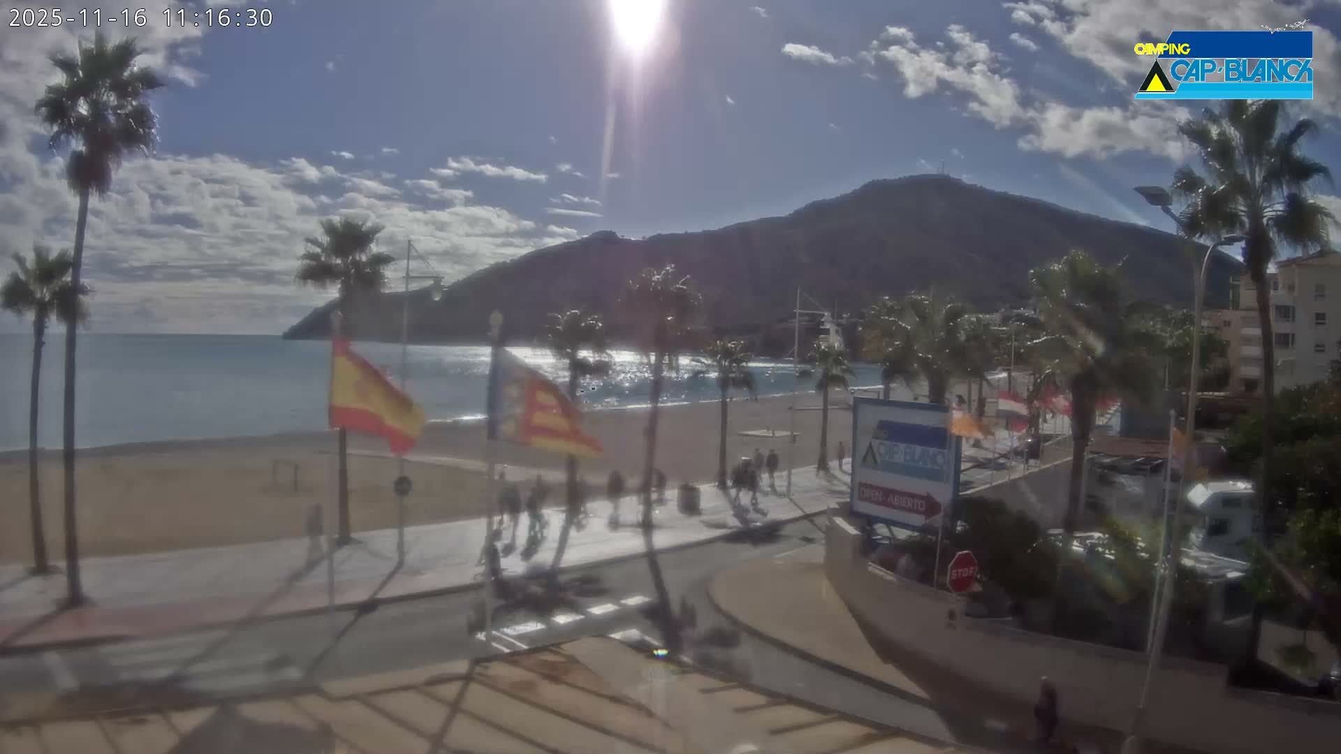 On a bright, sunny day with scattered clouds, a coastal scene unfolds with a sandy beach, calm sea, numerous palm trees, and a promenade bustling with pedestrians, all set against a backdrop of a large mountain.