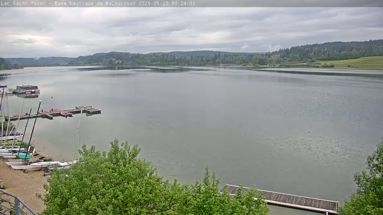 A calm lake under a cloudy sky is seen from a high vantage point, showing several small sailboats docked near a shoreline with green trees.