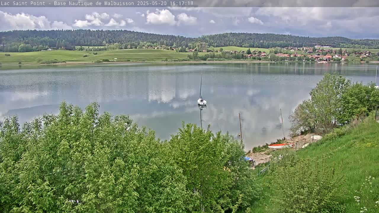 A calm lake reflects a cloudy sky, with a sailboat and several small boats visible near a shoreline partially obscured by lush green trees and grass.