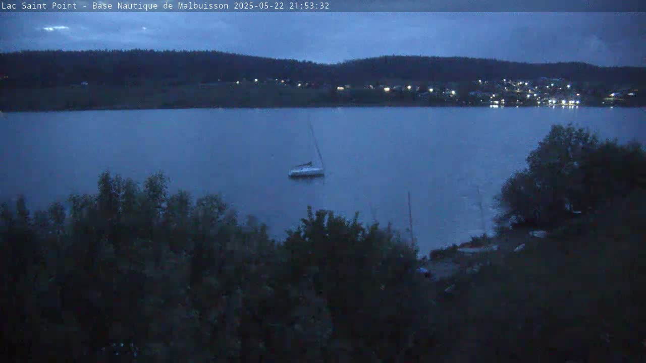 A sailboat sits still on a dark blue lake under a twilight sky, with trees and a distant town visible in the background.