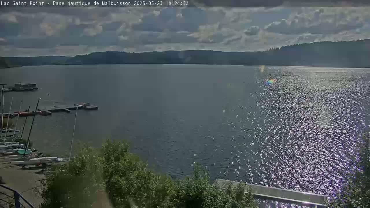A partly cloudy day shows a large body of water with sailboats docked near a shoreline, and a distant tree-lined hillside.