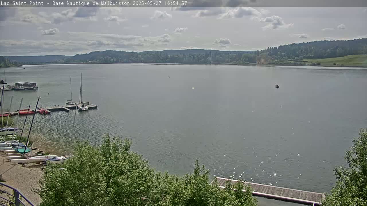 A calm lake with several sailboats docked near a shoreline, under a partly cloudy sky.