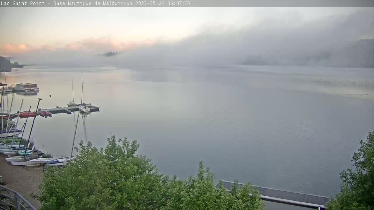 Several sailboats are moored at a dock on a foggy lake at sunrise.