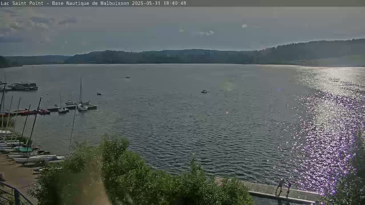 A calm lake with several sailboats docked near a shoreline, partially sunny with some clouds in the sky, and a few people visible in the lower right corner.