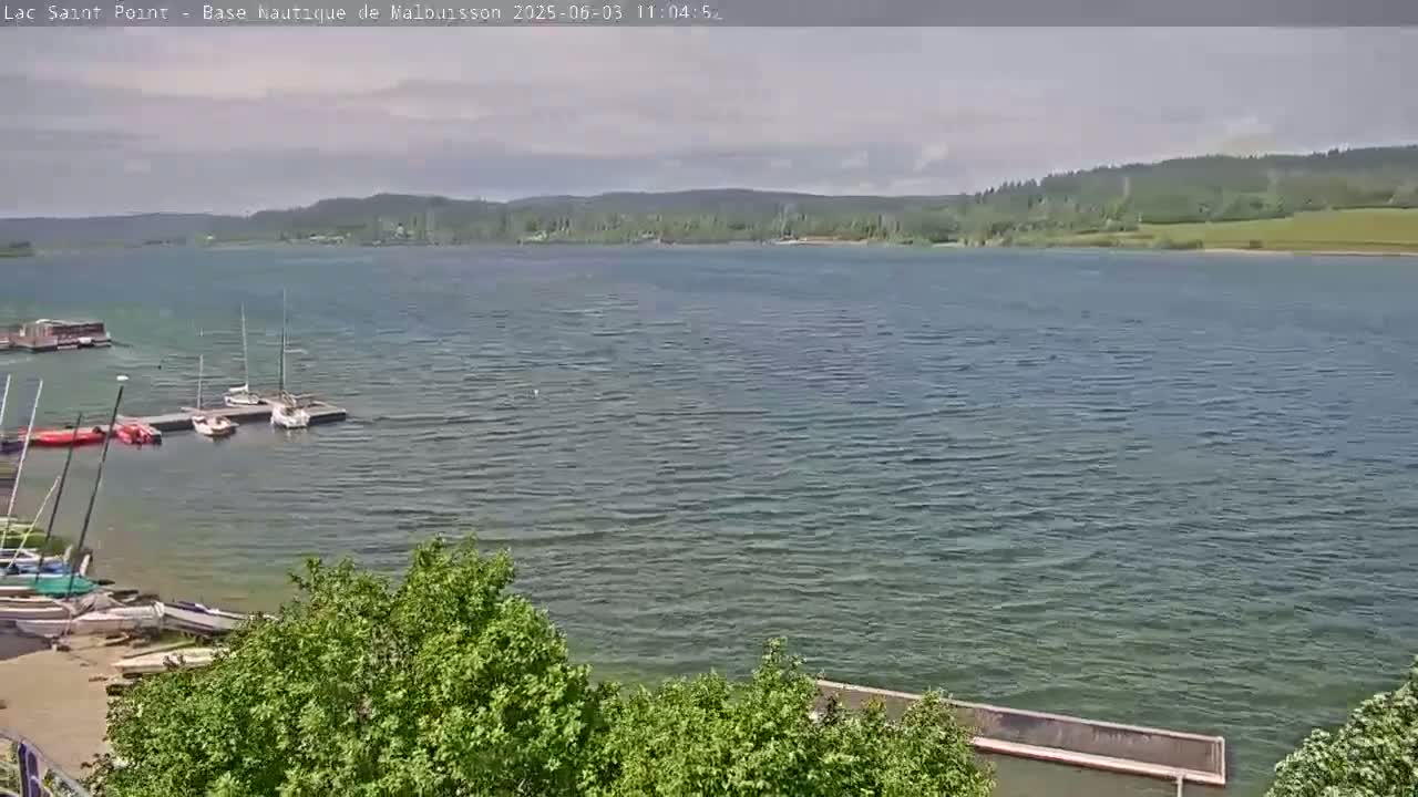 A partly cloudy day shows a calm lake with several sailboats docked near a shoreline with green vegetation and trees.