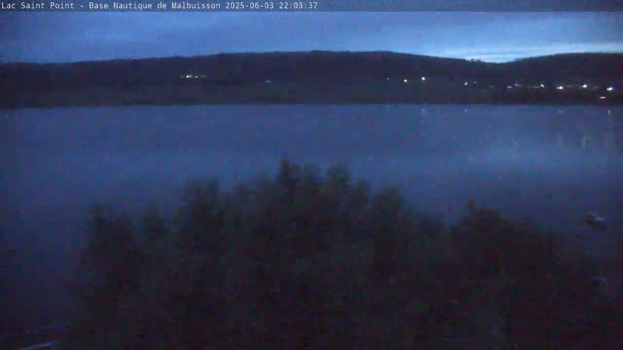 A dark, calm lake is seen at dusk, with a dark hillside in the background and vegetation in the foreground.