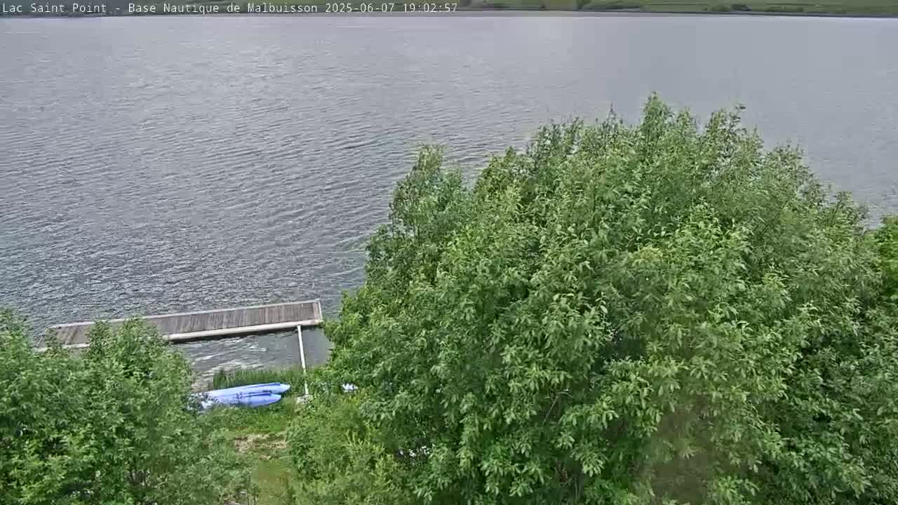 A calm body of water is seen from an elevated position, partially obscured by lush green trees and shrubs, with a wooden dock and a couple of small watercraft visible near the shore.