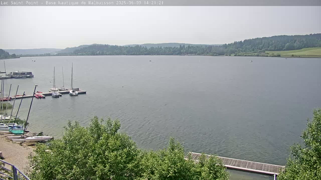 A calm lake with several sailboats moored at a dock,  under a hazy sky, is surrounded by green trees and hills.