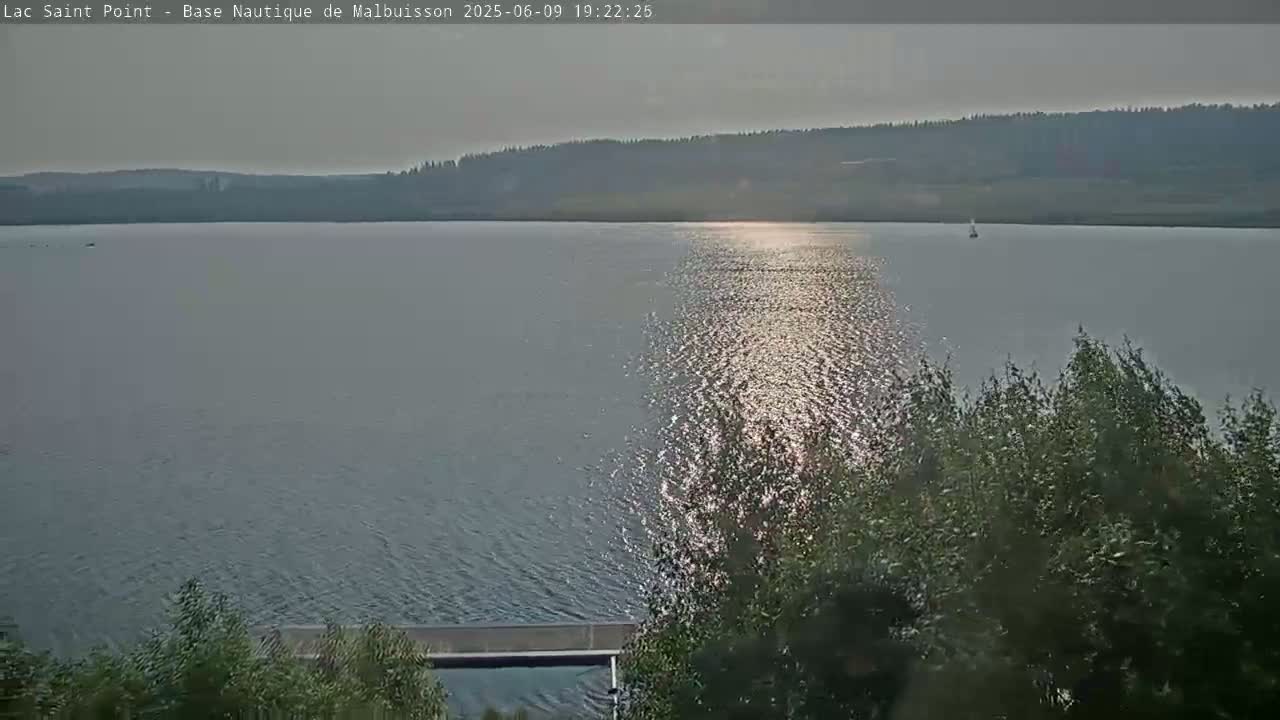 A calm lake reflects the sun's light, with a distant tree line visible under a hazy sky.