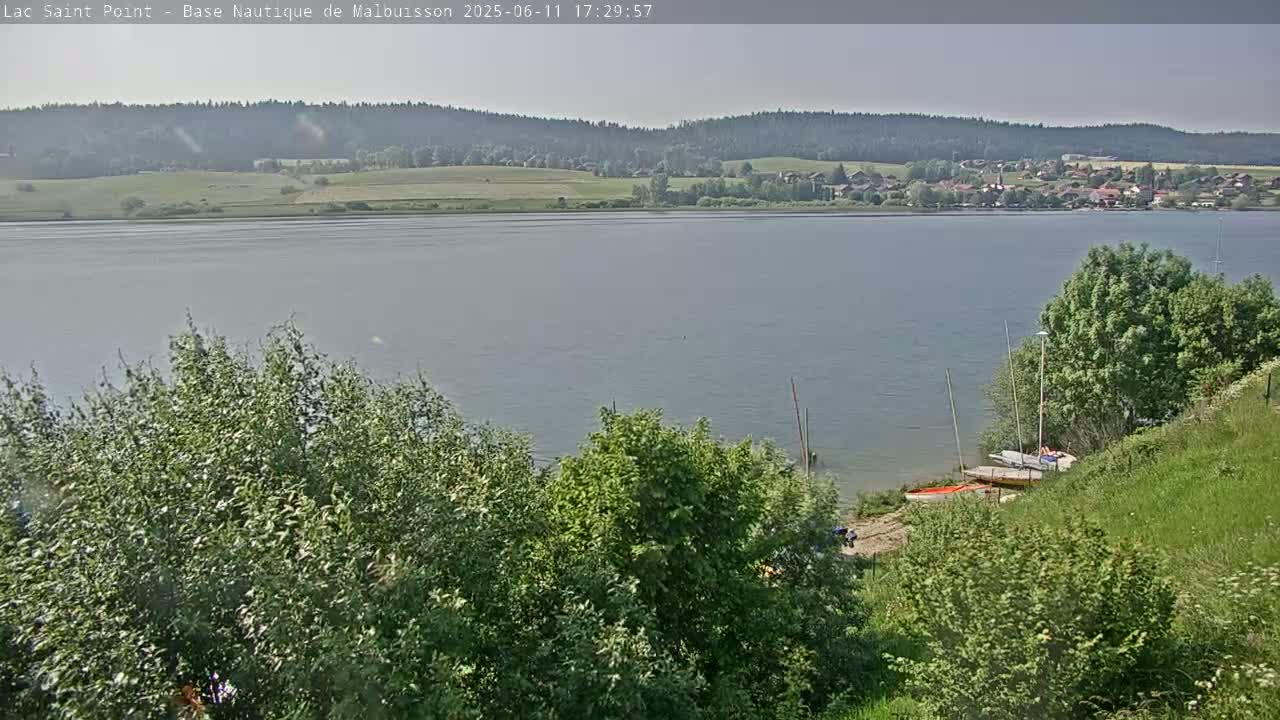 A calm lake is seen from a lakeside vantage point on a partly sunny day, with several sailboats beached near a grassy shoreline and a wooded area in the background.
