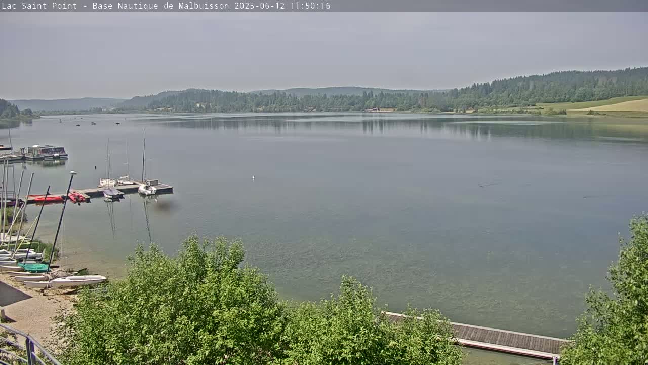 A calm lake with several sailboats and other watercraft moored near a shoreline with green trees under a hazy sky.