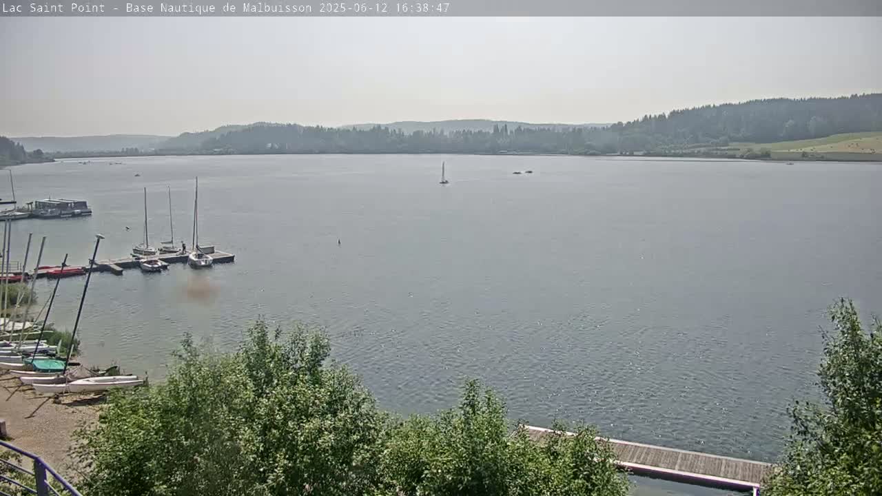 A calm lake with several sailboats moored at a dock, under a hazy sky.