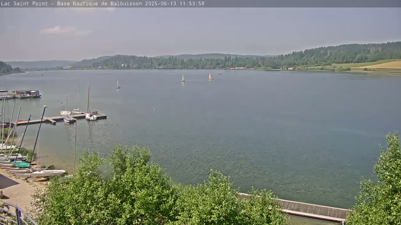 A calm lake is shown with several sailboats docked at a pier and two sailboats sailing in the distance on a hazy day.
