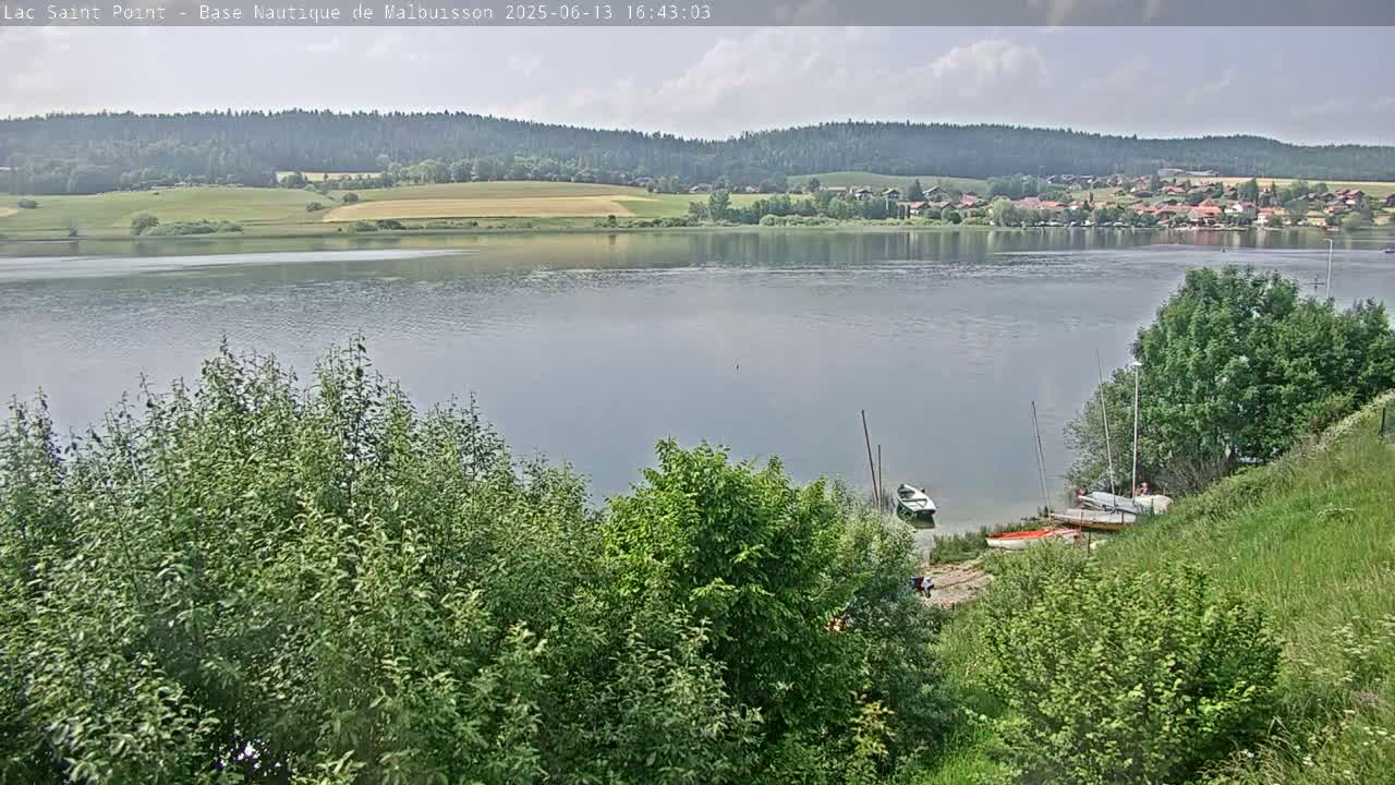A calm lake under a partly cloudy sky is surrounded by lush green trees and vegetation, with several small boats moored near the shore.