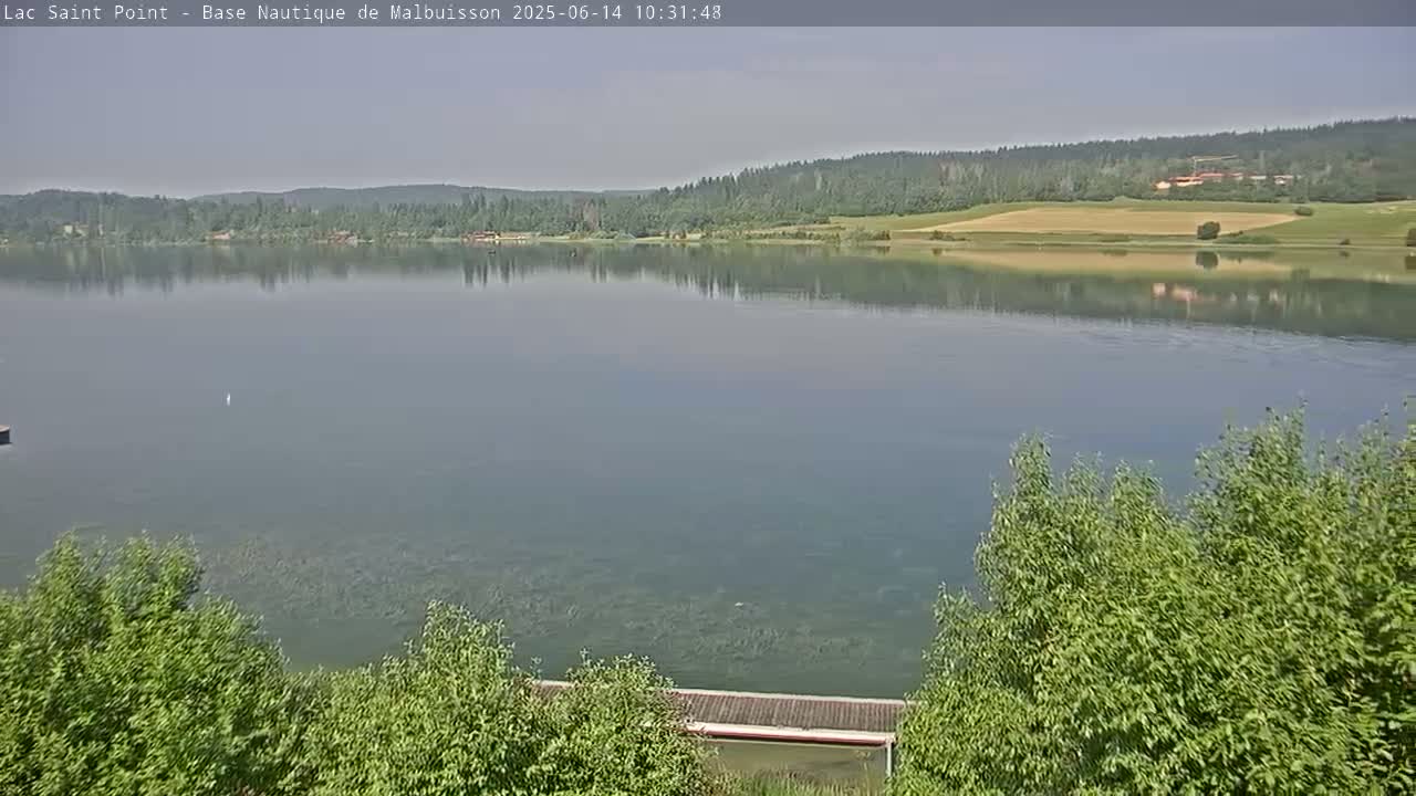 A calm lake reflects a tree-lined shore under a hazy sky.