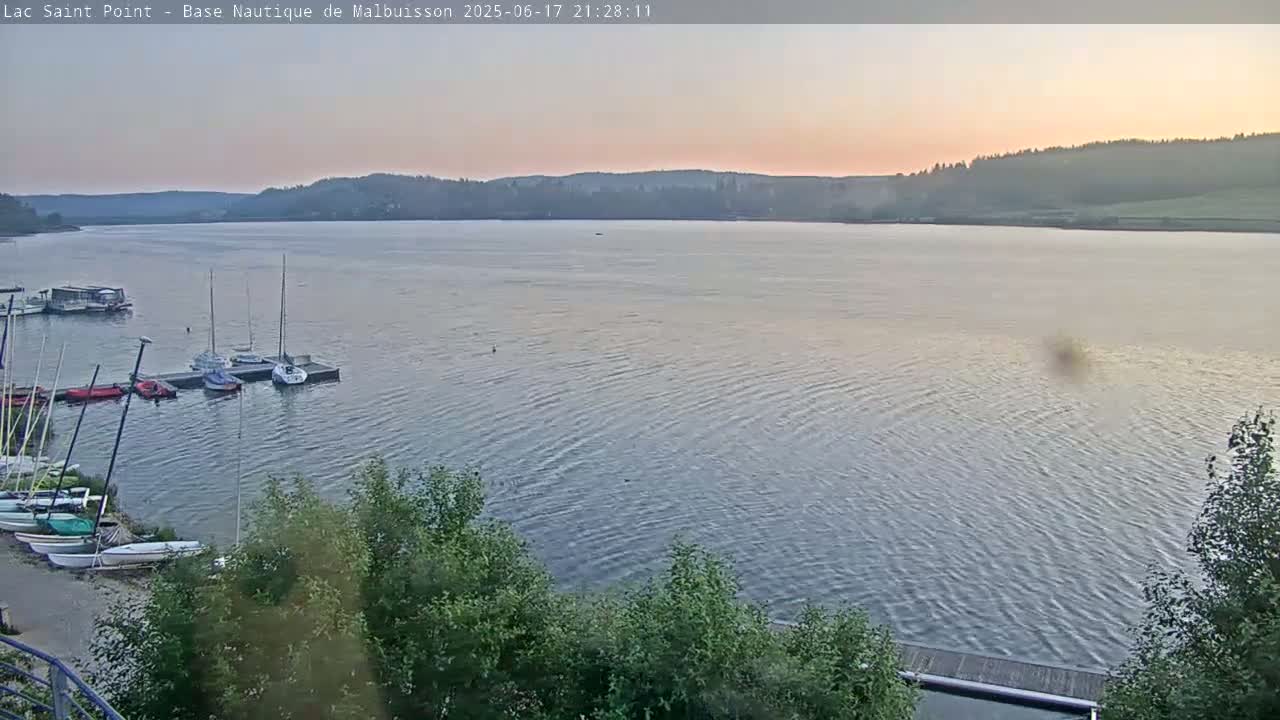 Several sailboats are moored at a dock on a calm lake at sunset, with a wooded shoreline in the background.