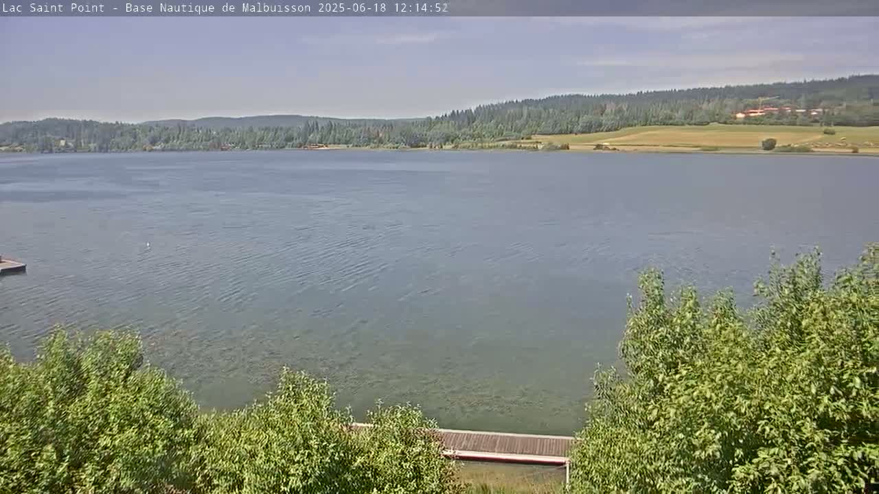 A calm lake is seen from a slightly elevated vantage point, with a forested shoreline in the distance under a mostly clear sky.