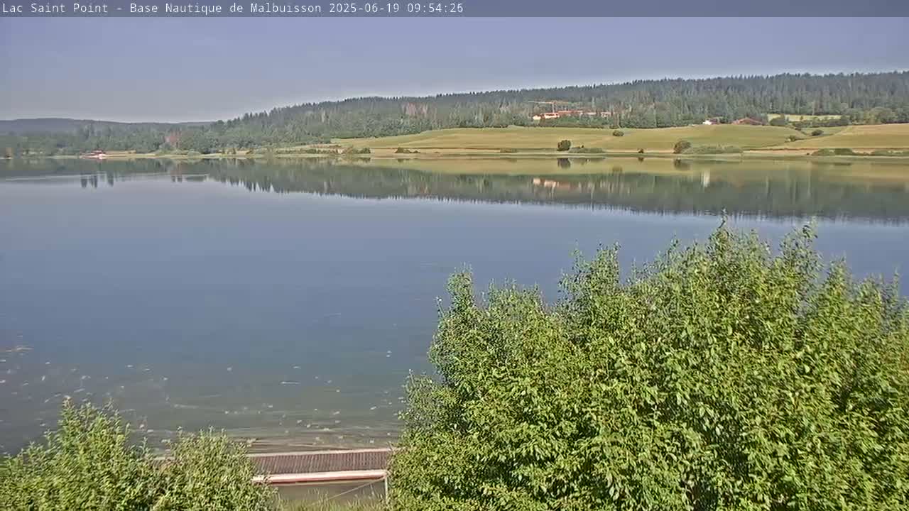 A calm lake reflects a treeline and grassy fields under a clear sky.