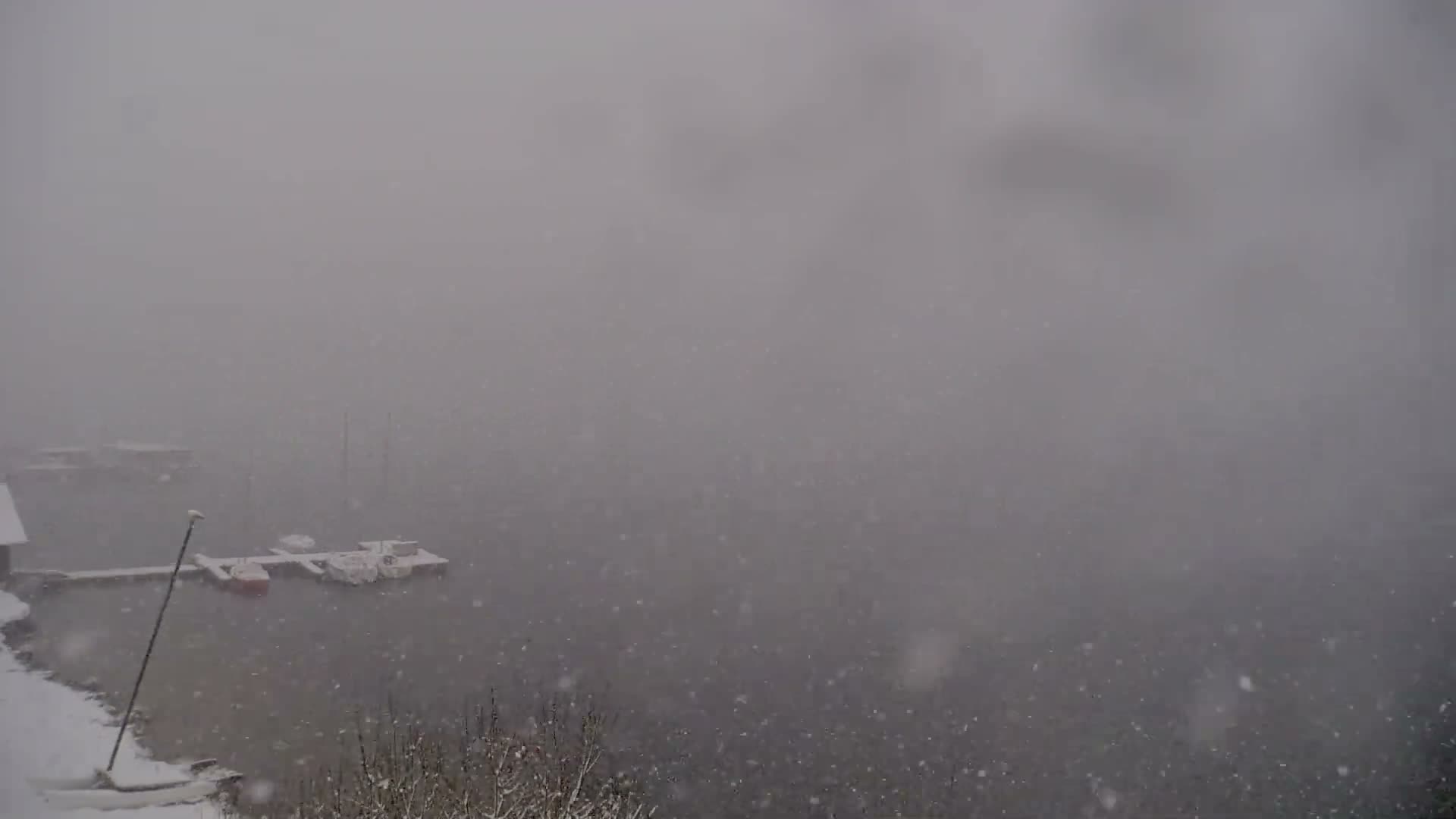 Heavy snowfall blankets a wintry scene featuring a body of water with a snow-covered dock mooring several small boats, while the distant landscape is obscured by falling snow and low visibility.