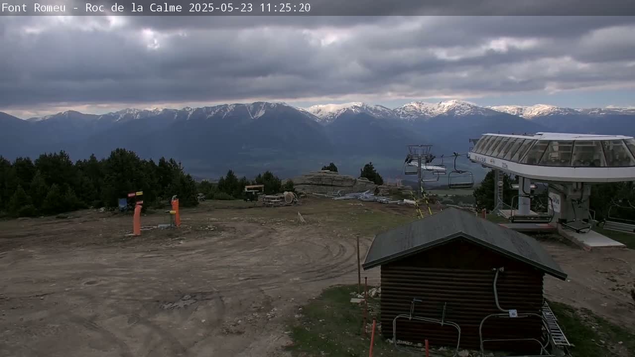 A mostly cloudy day reveals a mountain range capped with snow in the distance, a chairlift station with a small wooden building in the foreground, and a mostly barren, dirt area.