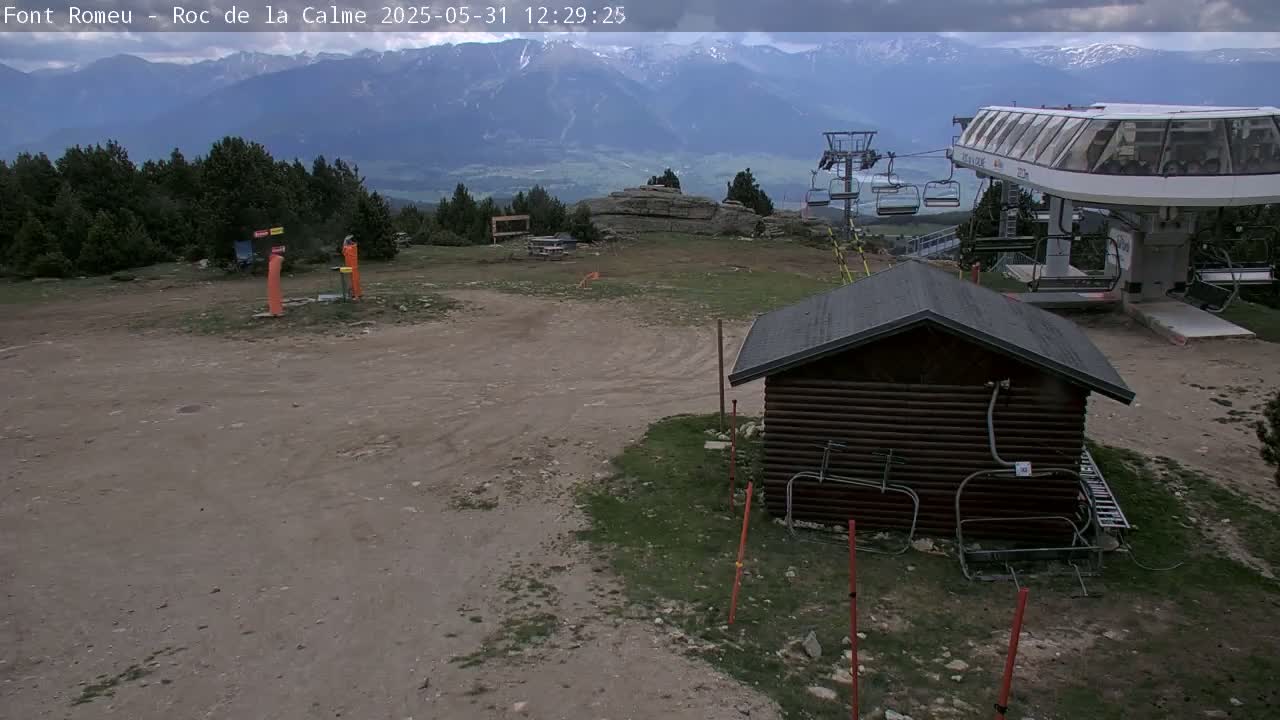 A mostly cloudy day at a mountaintop ski lift area shows a small wooden building, a chairlift, and a dirt path; mountains are visible in the distance.